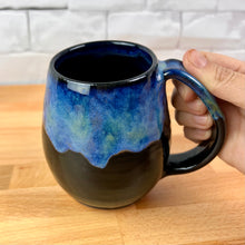 Load image into Gallery viewer, Aurora Borealis Mug. held by the artist, showing the generous handle. northwest style coffee mug thrown Red Stoneware, with large pulled handle. black glaze base with swirls of blue, turuoise, and hints of purple, reminiscent of the Aurora Borealis.