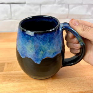 Aurora Borealis Mug. held by the artist, showing the generous handle. northwest style coffee mug thrown Red Stoneware, with large pulled handle. black glaze base with swirls of blue, turuoise, and hints of purple, reminiscent of the Aurora Borealis.