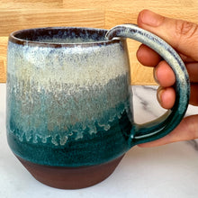 Load image into Gallery viewer, A ceramic mug with an angled shape, featuring a deep red-brown stoneware clay with a glossy glaze that transitions from white to teal. reminiscent of a forest view through the morning mist. A hand is holding the mug on a white marble surface with a light wood background. Forest Mist Mug. each one is an individual.