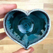 Load image into Gallery viewer, A wheel thrown and altered heart-shaped ceramic bowl with a glossy teal and white dripping glaze, held by a hand over a light wood background.