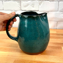 Load image into Gallery viewer, Teal ceramic pitcher held by a hand on a wooden surface with a white tiled wall background. Wheel thrown pottery pitcher shown in Teal with pulled handle. handcrafted and wheel thrown at  Fern Street Pottery
