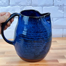 Load image into Gallery viewer, Blue ceramic pitcher held by a hand on a wooden surface with a tiled wall background. Wheel thrown pottery pitcher shown in Blue with pulled handle. handcrafted and wheel thrown at  Fern Street Pottery