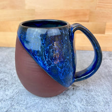 Load image into Gallery viewer, One of a kind glaze. Handcrafted, wheel thrown mug with pulled handle. Blue ceramic mug with a mottled blue glaze held by a hand on a concrete surface.