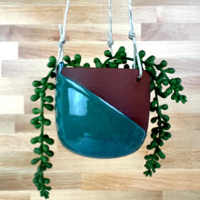 Load image into Gallery viewer, A hanging ceramic planter with a diagonal split design in glossy moss green glaze and dark reddish-brown stoneware, suspended by hemp twine. shown with trailing green succulent stems.