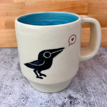 Load image into Gallery viewer, artisan made, wheel thrown pottery mug. porcelaina clay, with a cute crow (or raven) image that is both painted on and carved into it. the crow speaks of love as it is speaking a heart in a speech bubble. glossy Sky Blue interior glaze. 