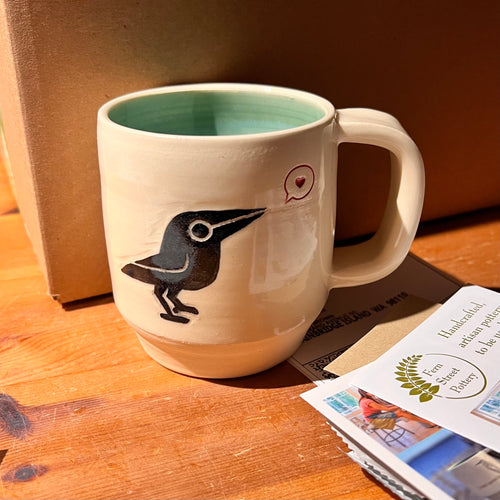 artisan made, wheel thrown pottery mug. porcelaina clay, with a cute crow (or raven) image that is both painted on and carved into it. the crow speaks of love as it is speaking a heart in a speech bubble. glossy turquoise interior glaze. 