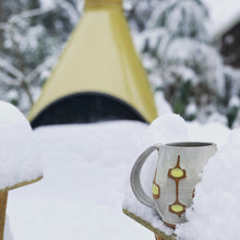 Load image into Gallery viewer, wheelthrown Pottery mug, hand glazed with MidMod pattern in white, orange and yellow, shown in a snow scene in front of a Mid Century style fireplace, in a snowy scene. Fern Street Pottery