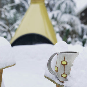 wheelthrown Pottery mug, hand glazed with MidMod pattern in white, orange and yellow, shown in a snow scene in front of a Mid Century style fireplace, in a snowy scene. Fern Street Pottery