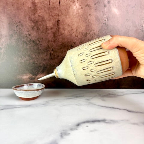 Olive oil cruet. Wheel thrown and carved. cork stopper  for smooth pouring. Thrown in a red stoneware clay and carved by hand. the white glaze accentuates the carved pattern where the red clay shows through. shown pouring into a tiny sauce bowl.