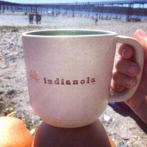 Wheel thrown pottery mug with "indianola" and an image of a crab inset on the outside. white outside, turquoise green glaze interior