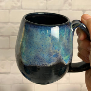 Aurora Borealis mug, blue over black glaze, northwest style coffee mug thrown pottery, with large pulled handle. shown held by the artist