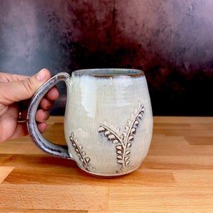 a wheel thrown and hand carved mug is held by the artists. the mug is thrownin red stoneware which shows throught the white glaze at the edges of the mug. the mug has a delicate vine pattern carved into it.