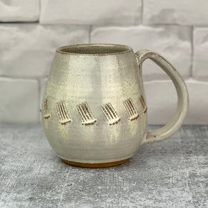 a wheel thrown and hand carved mug sits in a kitchen setting. the mug is thrownin red stoneware which shows throught the white glaze at the edges of the mug. the mug has a repeated line pattern scratched pattern carved into it.